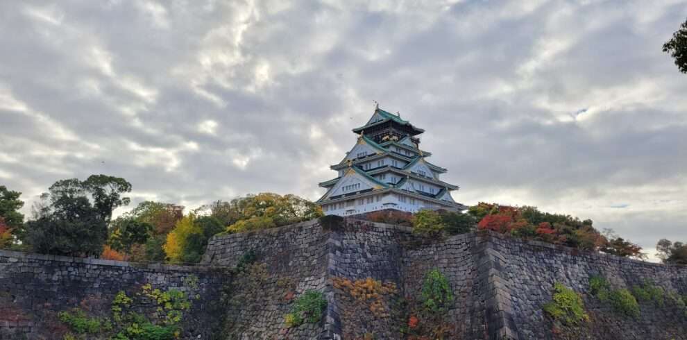Famous white gold and blue Osaka Castle behind its walls covered in autumn colours and a cloudy sky