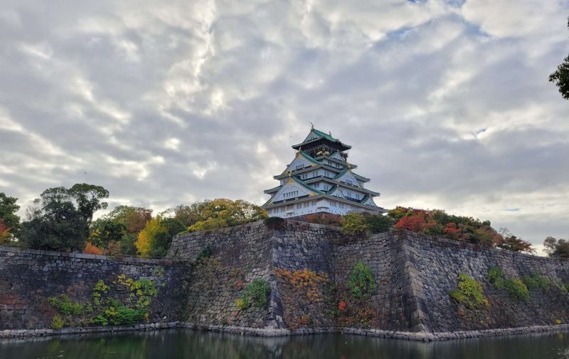 Famous white gold and blue Osaka Castle behind its walls covered in autumn colours and a cloudy sky